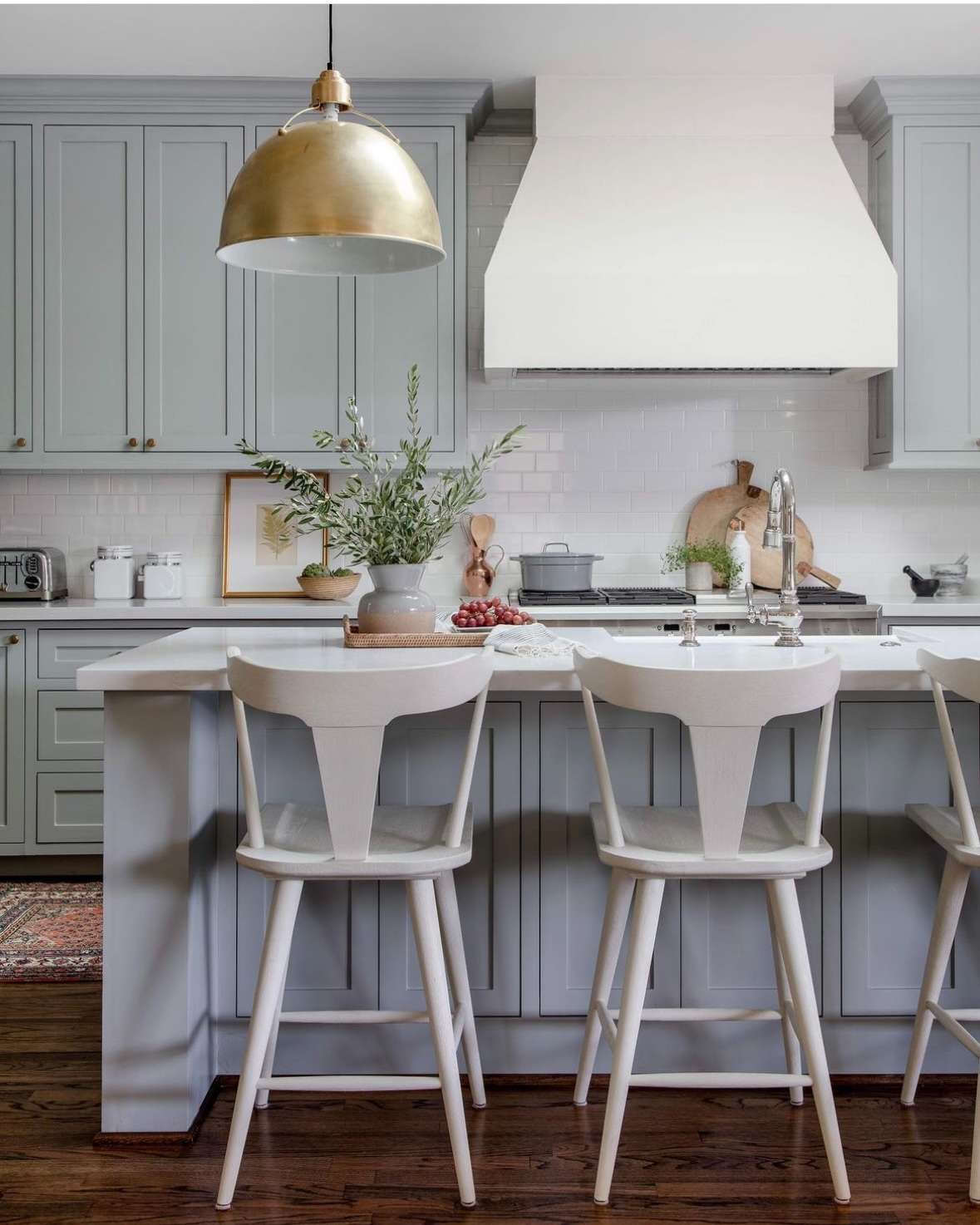 A kitchen with sage green cabinets, white marble countertops, white subway tile backsplash, and a large gold pendant light.