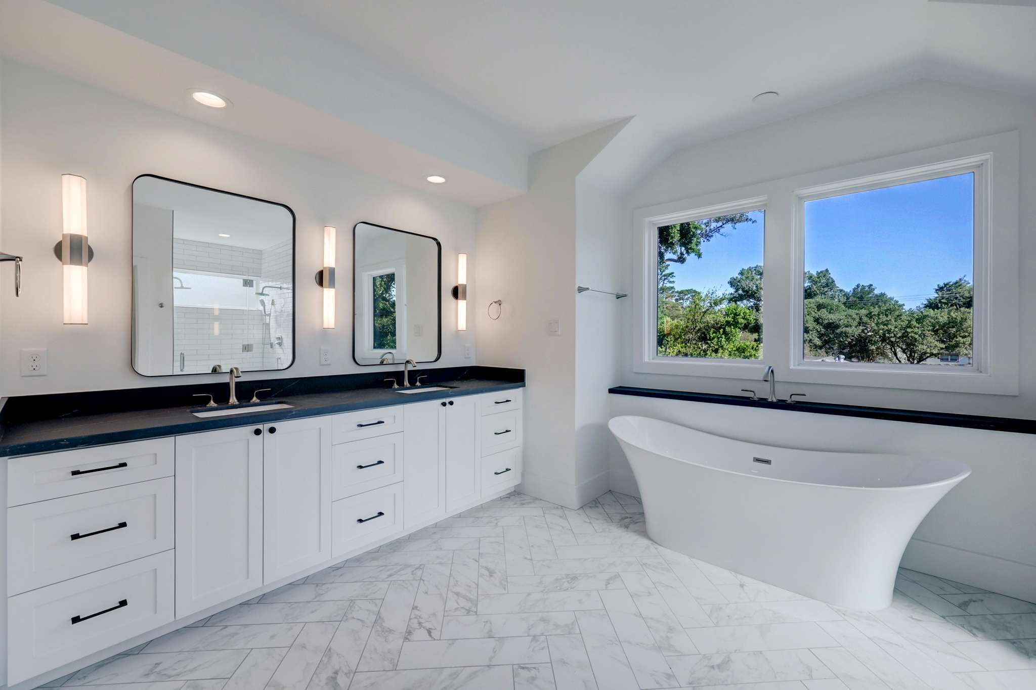 A bright bathroom with a white freestanding tub near a window, dual vanity with black countertops, and white cabinets.